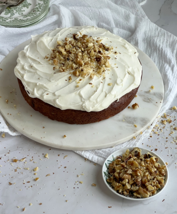 old fashioned banana cake in a Garten on a piece of marble with a bowl of walnuts next to it and a pile of green and white transfer ware plates and forks.