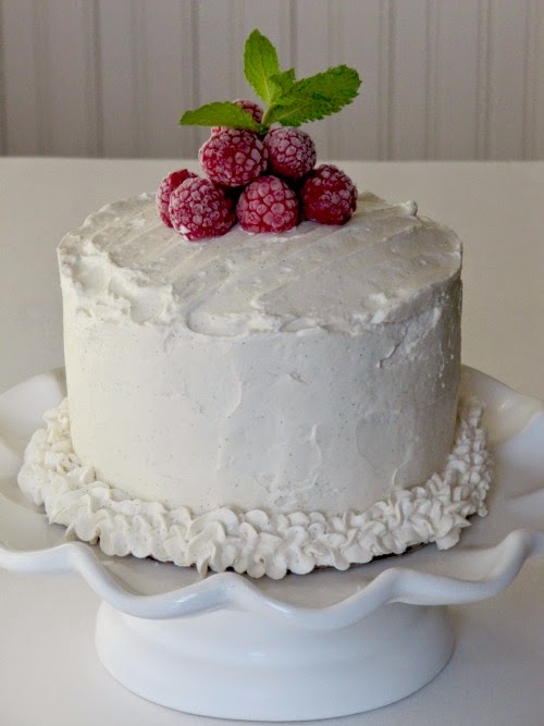 A cake frosted with white icing topped with raspberries sitting on a white cake platter with a ruffled edge.