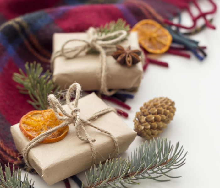Christmas gifts wrapped in brown paper sitting on a plaid blanket with oranges and pinecones.