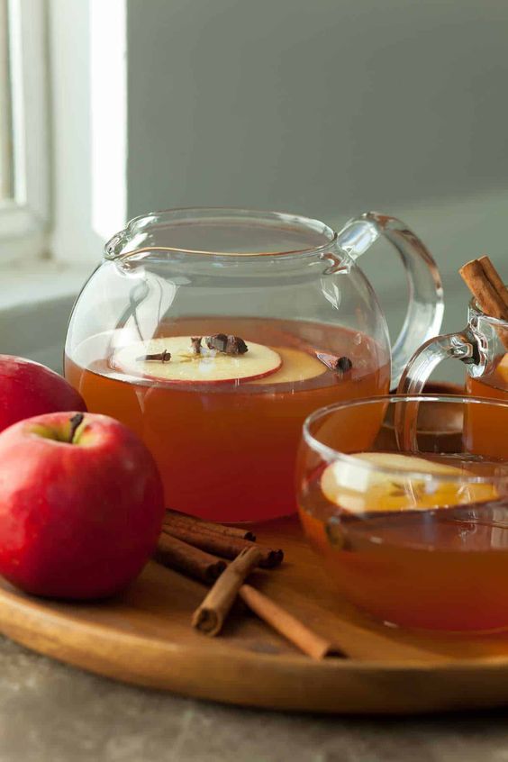 a clear glass pitcher filled with apple tea and a red apple sitting next to it and a glass of tea