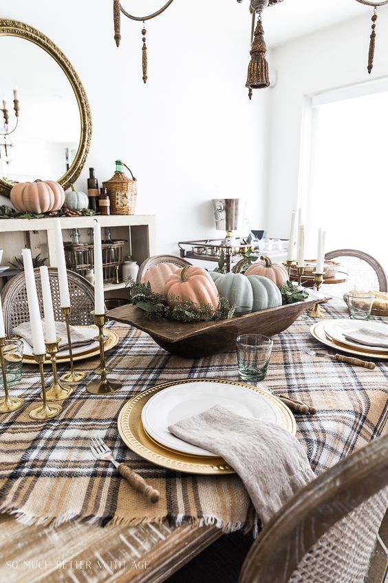 ding table decorated for thanksgiving with plaid table cloths, a large dough bowl in the center filled with pumpkins and set for dinner with gold chargers and white dinner plates