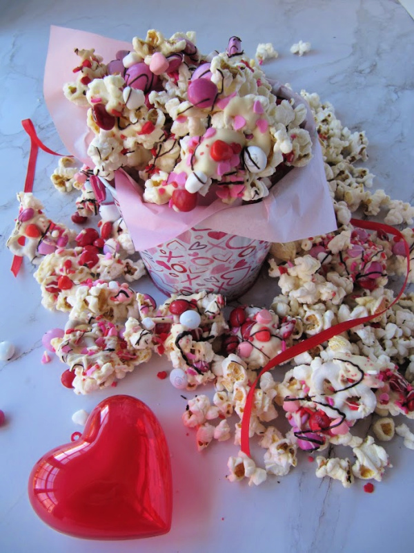 Bucket of chocolate covered popcorn with colored candy and sprinkles and a red plastic heart sitting next to it.