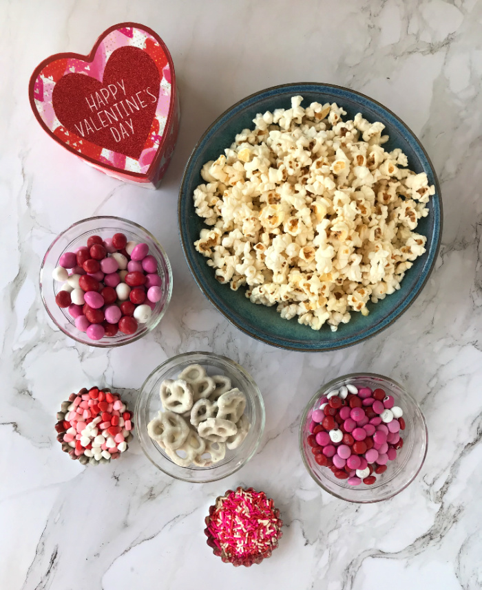 Blue Bowl of Popcorn surrounded by small bowls of colored candy, sprinkles, jimmies and pretzels