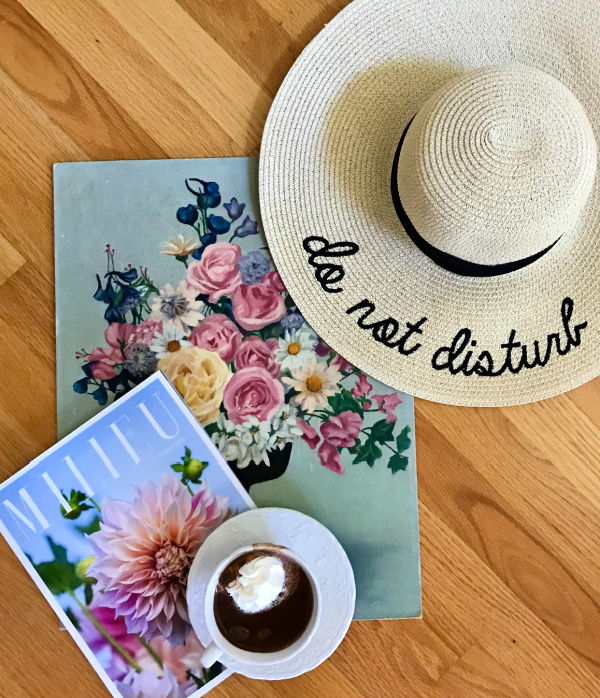 Do not disturb sun hat sitting on top of a flower painting with a milieu magazine and a cup of hot chocolate