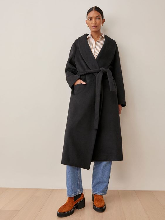 Woman leaning against a wall wearing jeans brown shoes and a black coat tied at the waist
