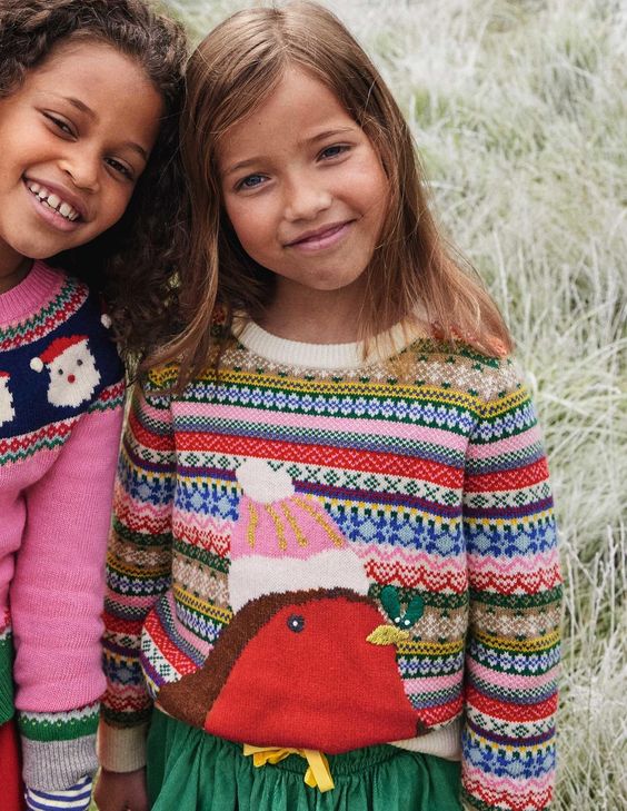 Two little girls wearing christmas sweaters, the one on the left is wearing a pink sweater with Santas and the little girl on the right is wearing a fair else sweater with a robin