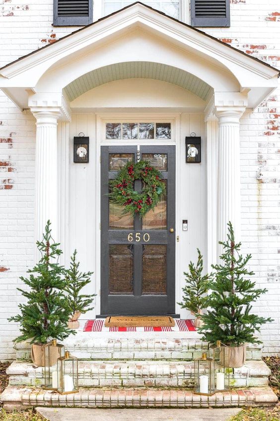 White distressed brick house with black door that has a green wreath and 4 little christmas trees.