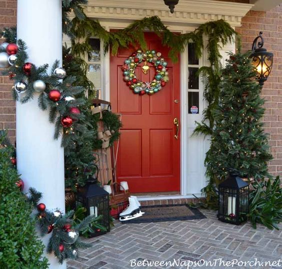Red door with a wreath and a christmas tree on the right of the door, white columns wrapped in pine garland and a pair of ice skates to the left.