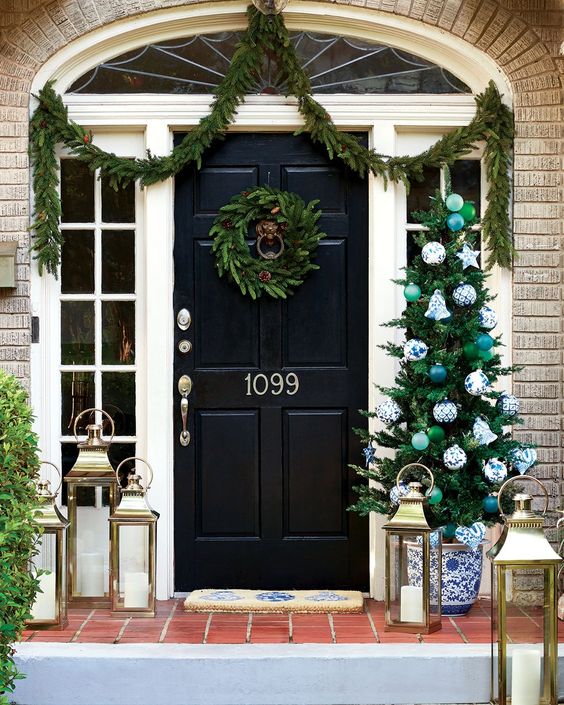 brick house with black door and a Christmas wreath a swag of greens and a christmas tree and lanterns.