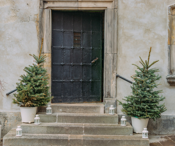 two Christmas trees in white planters on the steps of an old building with white lanterns next to them
