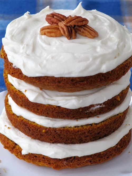 a four layer cake with white frosting with pecans on the top sitting on a white plate and a blue plaid tablecloth