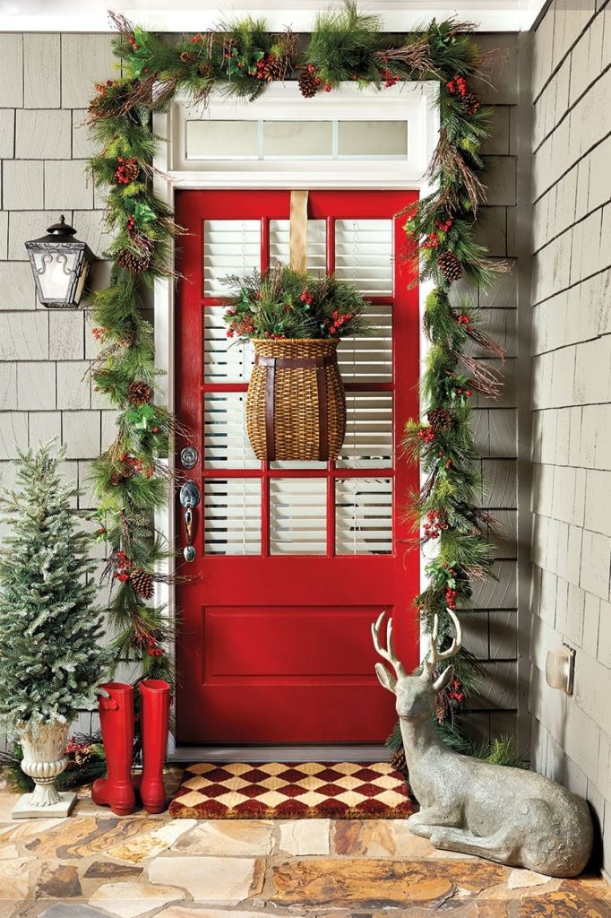 Red door with a wicket basket filled with greens. A garland around the door and a pair of small chrstmas trees on the leftt go the door with a pair of red boots and a small deer on the right side, front door decorating for christmas. 