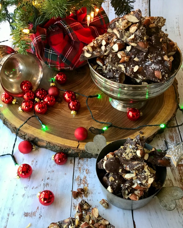 caramelized almond bark in a silver-plate bowl sitting on a wood slab with a small bowl next to it. The wood slab and the backroad are decorated with christmas lights and ornaments