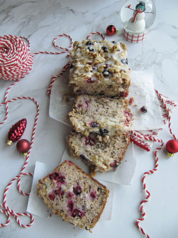 cranberry walnut loaf wrapped in white paper and tied with red and white string on a white piece of marble with scattered christmas bulbs a snow globe and string