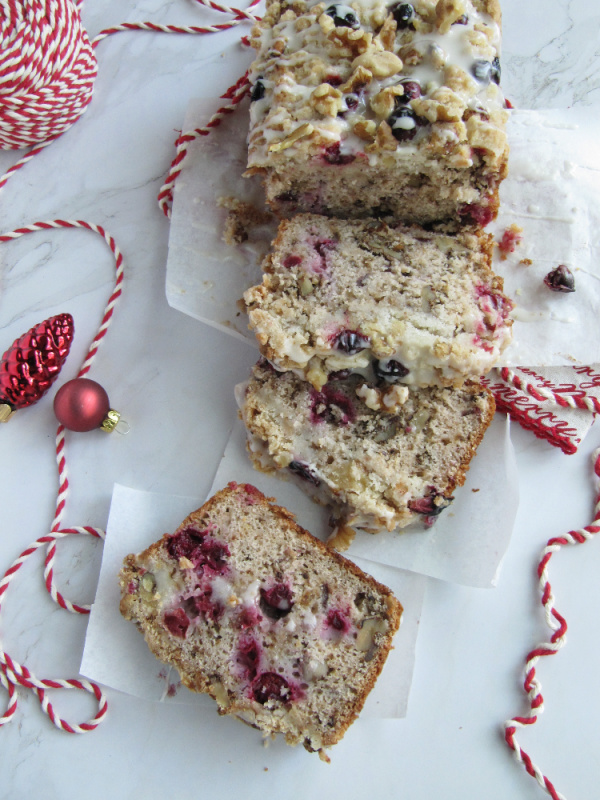 cranberry walnut loaf wrapped in white paper and tied with red and white string on a white piece of marble with scattered christmas bulbs a snow globe and string