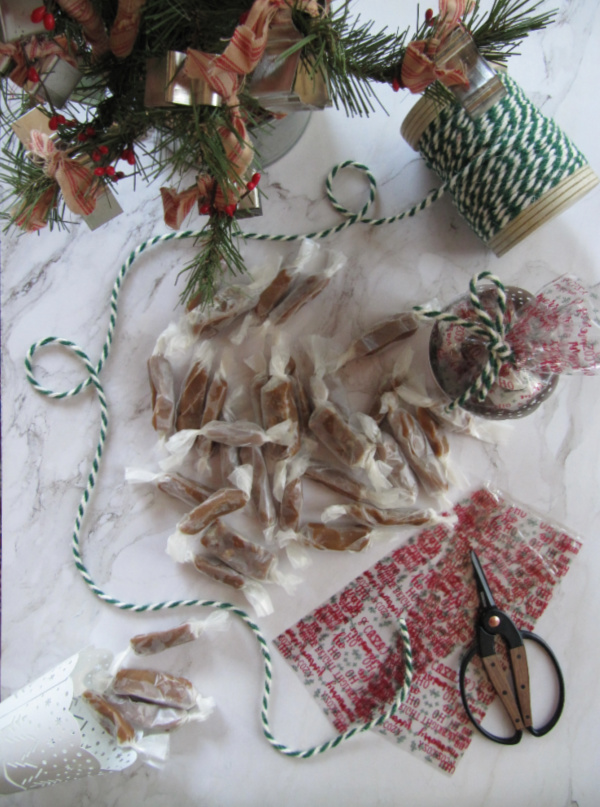 flatly on white marble with a christmas tree green and white twine cellophane bags scissors and homemade caramels wrapped in waxed paper, holiday hostess gifts. 