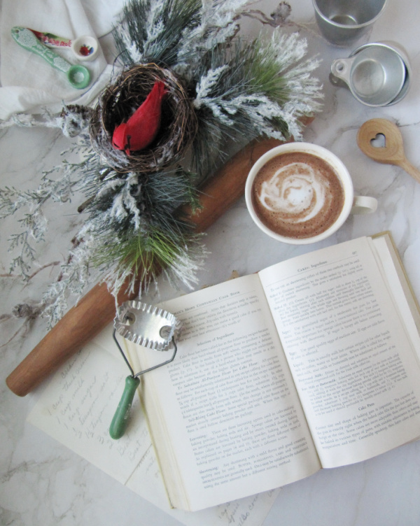 Flatlay with vintage cookbook, handwritten recipe, cup of hot chocolate, evergreen and a nest with a cardinal and various vintage cooking tools