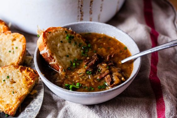 white bowl of short rib onion soup with a spoon on the right and a piece of toasted bread on the left