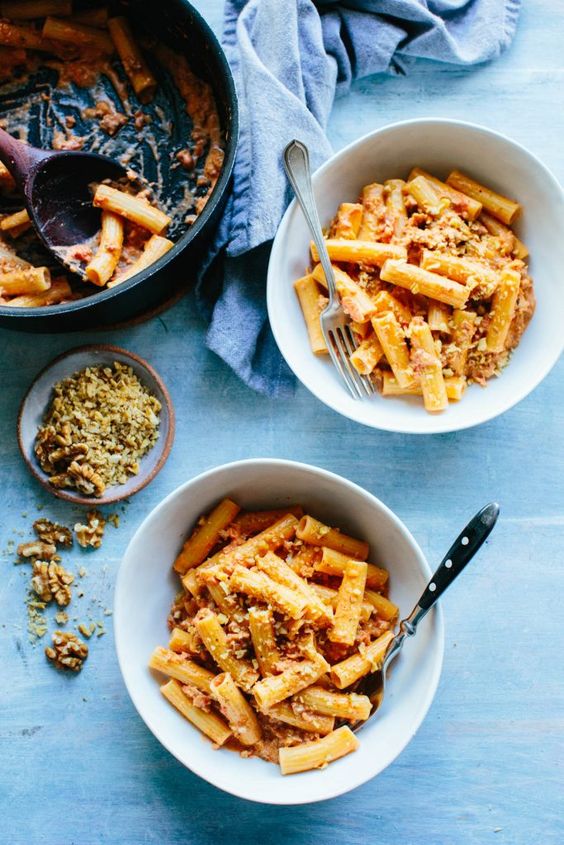 Two white bowls filled with sausage rigatoni pasta with a skillet and a bowl of walnuts on a blue background