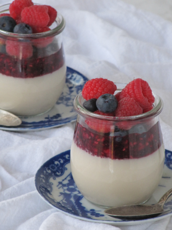almond milk panna cotta in 2 clear jars with berries sitting on blue and white transfer ware plates. 