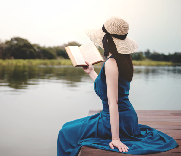 woman wearing a blue dress and a straw hat sitting on the edge of a wood pier reading a book by water