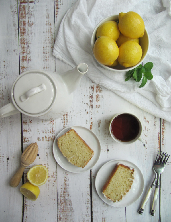 Lemon loaf cake on white plates with a pot of tea and a bowl of lemons