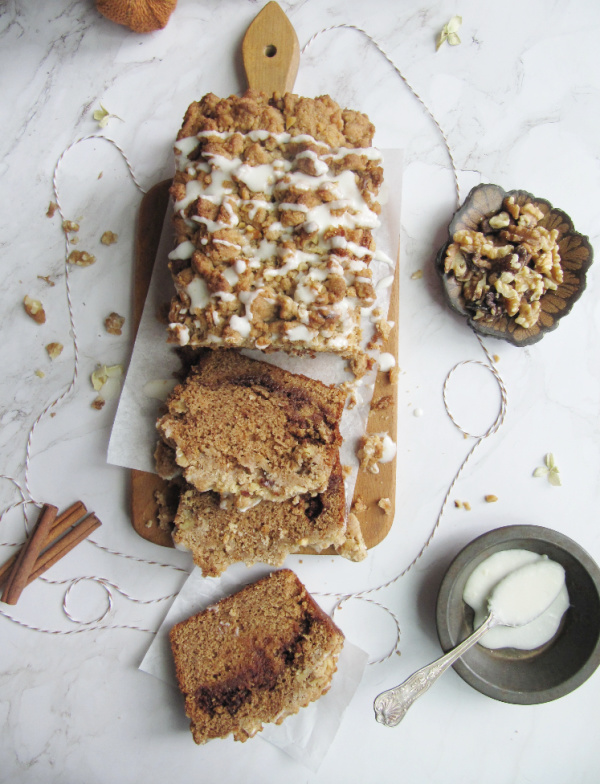 cinnamon swirl pumpkin bread with crumble topping on a wood cutting board with a bowl of walnut on the left and some frosting