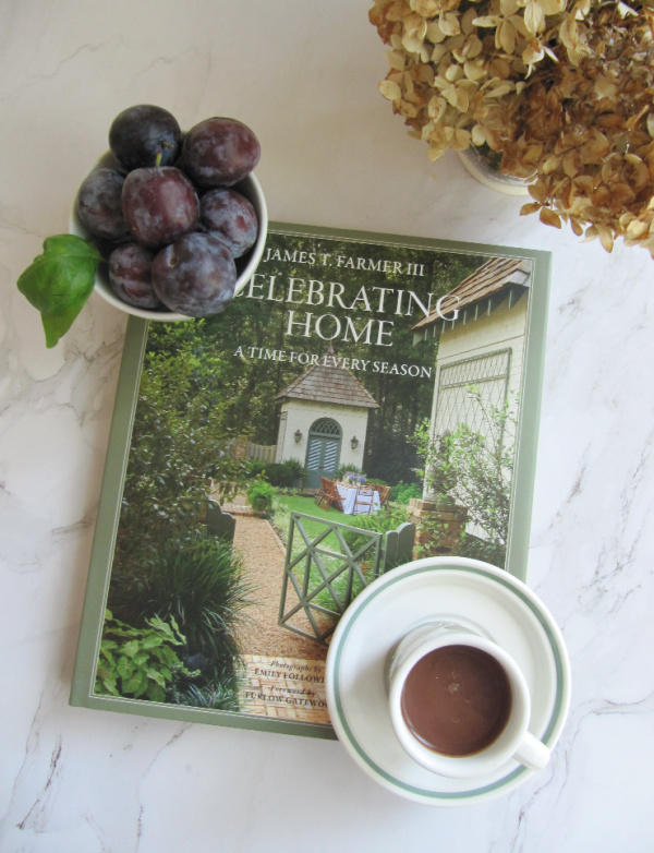flatlay James farmer book bowl of plumbs in the left corner and dried hydragneas with a cup of hot chocolate.