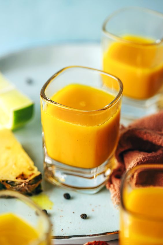 4 shot glasses filled with orange liquid on a tray with slices of lime and pineapple. 