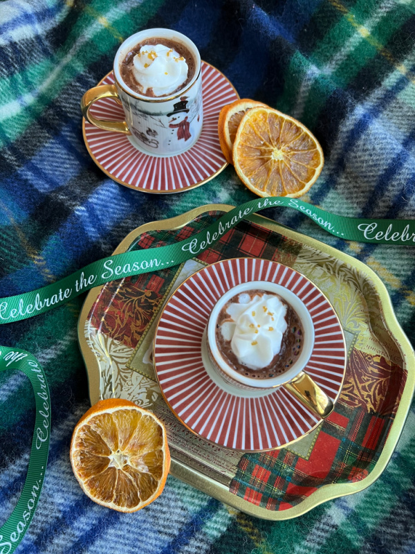 2 small cups of hot chocolate with whipped cream on a tray sitting on a plaid blanket with dried oranges.