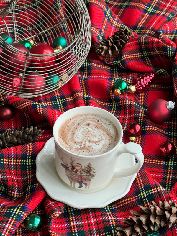 flatlay with wire basket filled with christmas bulbs on a plaid blanket with a cup of hot chocolate in an ivory mug with a vintage Santa and reindeer.