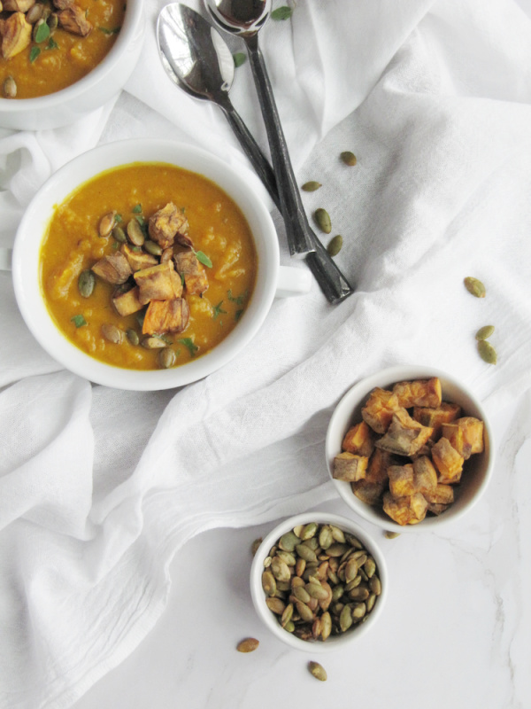 Soup in white bowls with 2 spoons and two small bowls of potatoes and pumpkin seeds. 