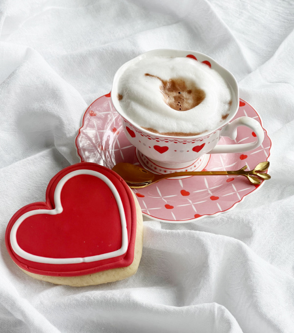 cup of hot chocolate with foam in a white cup with hearts on a pink saucer with a heart cookie.