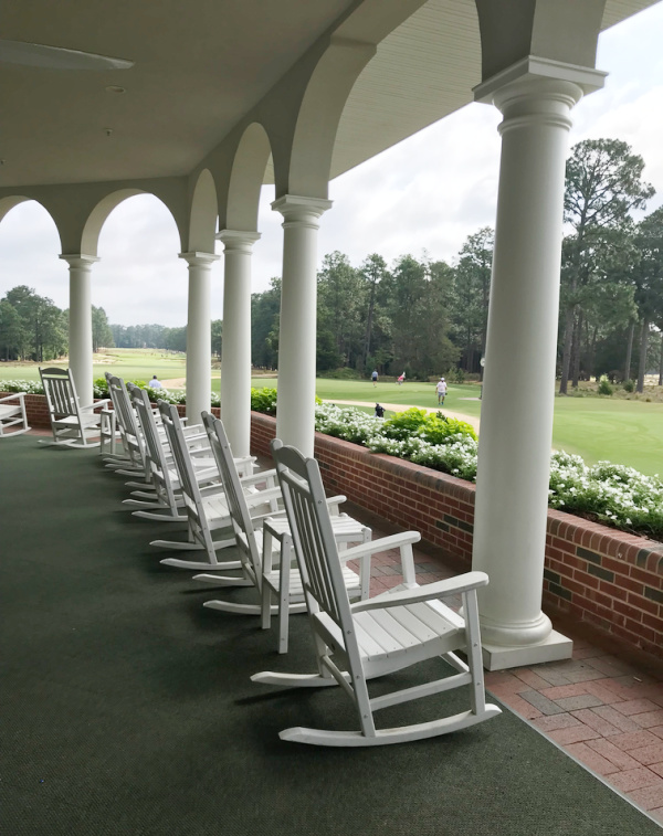 A row of white rocking chairs on a porch overlooking a golf green.