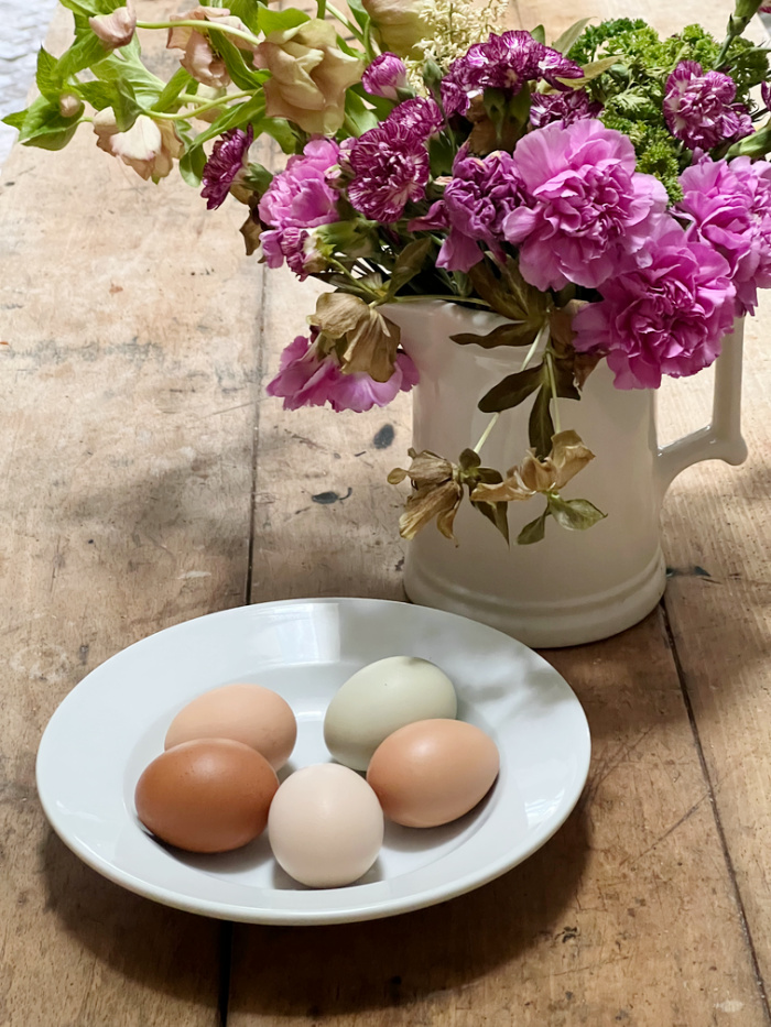 farm raised eggs pf various colors in a white bowl and a pitcher of flowers.