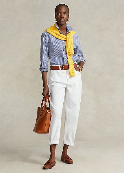 Woman wearing a pair of white jeans, blue and white stripe shirt and a yellow sweater with a brown handbag. 