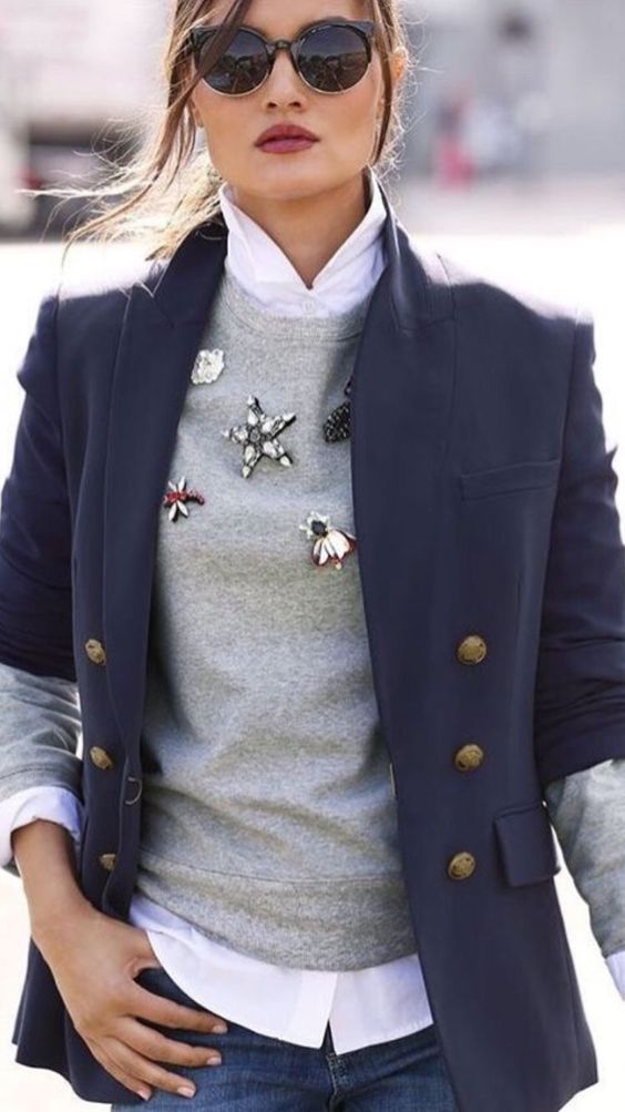 Woman dressed in a pair of jeans, a white blouse, gray sweater with brooches and a blue blazer. 