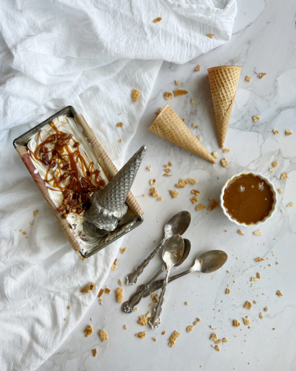 Salted Caramel Candied Pecan "No Churn" Ice Cream in a loaf pan with a bowl of caramel and cones laying by the side.