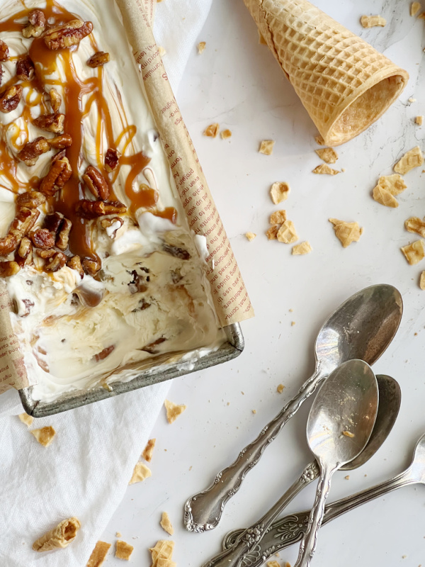 Salted Caramel Candied Pecan "No Churn" Ice Cream in a loaf pan with 3 spoons and a sugar cone next to it.