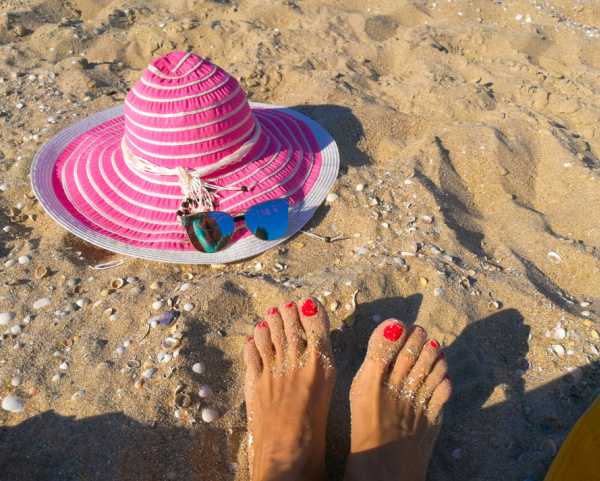 A pink sun hat laying in the sand near 2 feet with painted red toenails.