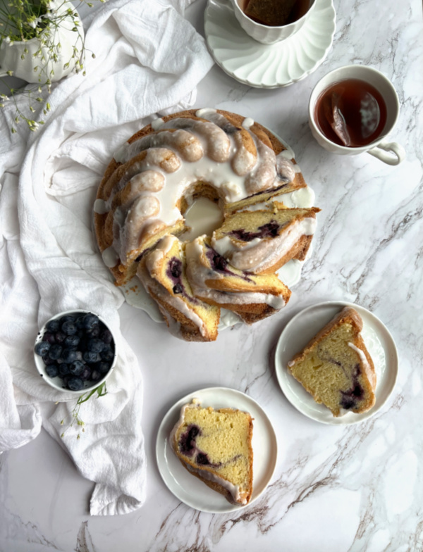 blueberry swirl bundt cake cut on a platter with 2 pieces on white plates and a cup of tea and extra blueberries nearby.