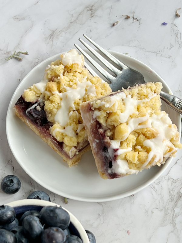 blueberry crumb bars stacked in a plate with a fork and a small bowl of blueberries.