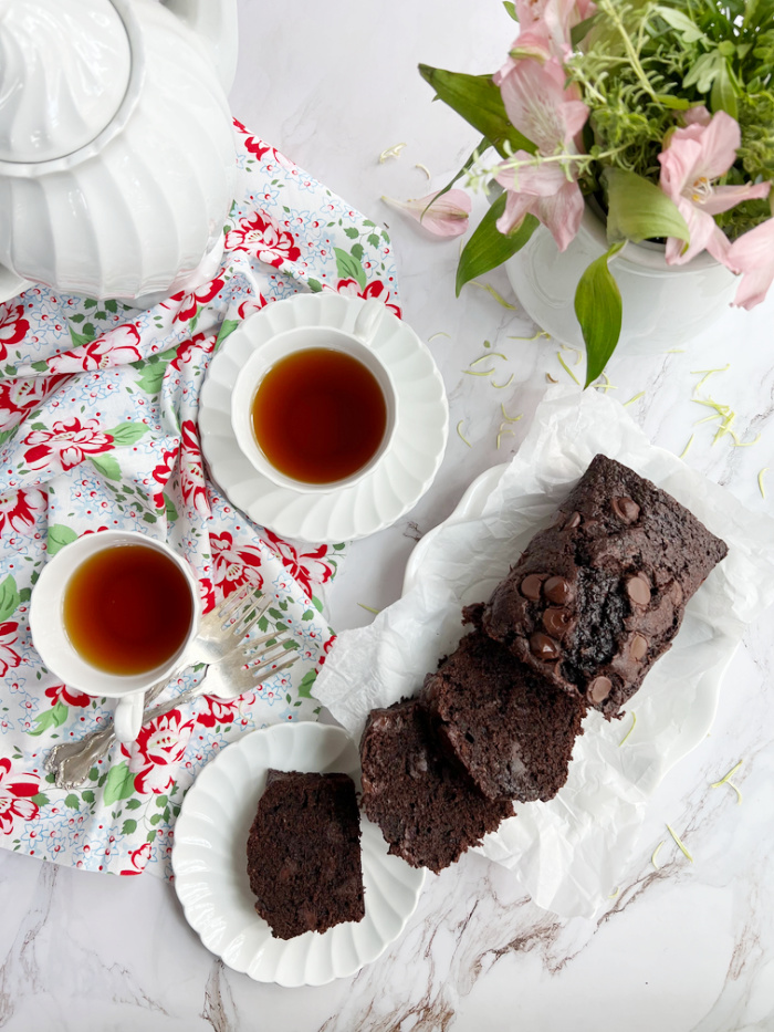 double chocolate chip zucchini bread sliced on a white plate with 2 cups of tea a white teapot and a vase of flowers. 