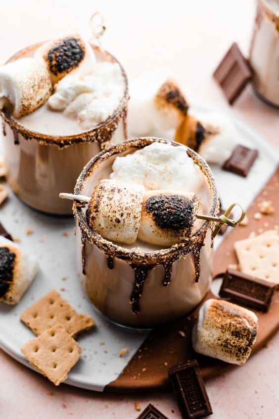 S'mores cocktails, 2 with broken graham crackers, chocolate and marshmallows scattered around. 