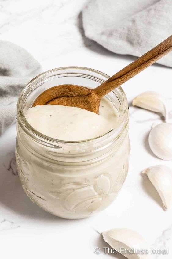 White Garlic Pizza Sauce in a glass jar with a wood scoop sticking out the top and peeled garlic laying around it. 