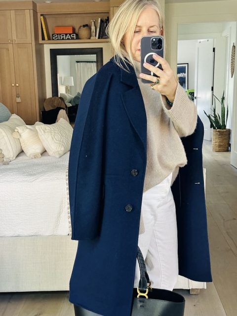 Woman wearing a pair of white pants, tan sweater, blue coat and black bag. 