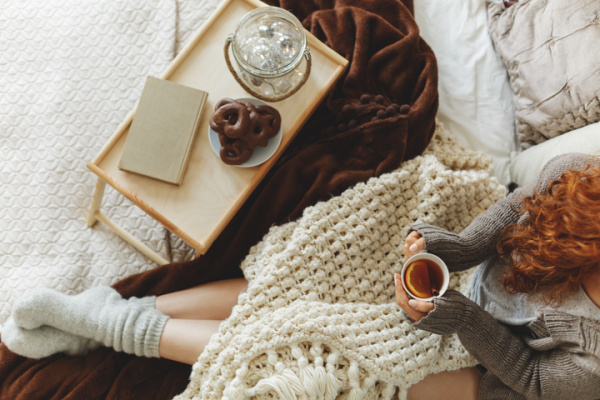 15 Tips for a Healthier Life A girl with a cup of tea curled up in a knitted blanket in bed with a book and plate of chocolate pretzels.