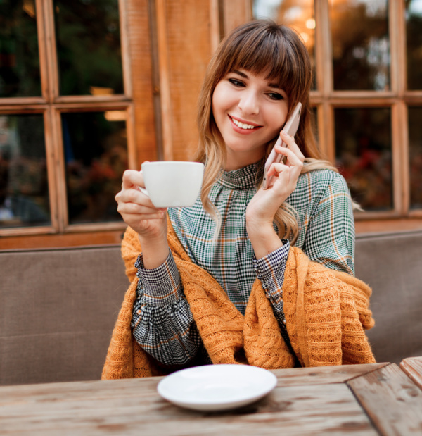 Woman wearing a plaid dress drinking coffee and talking on the phone.
