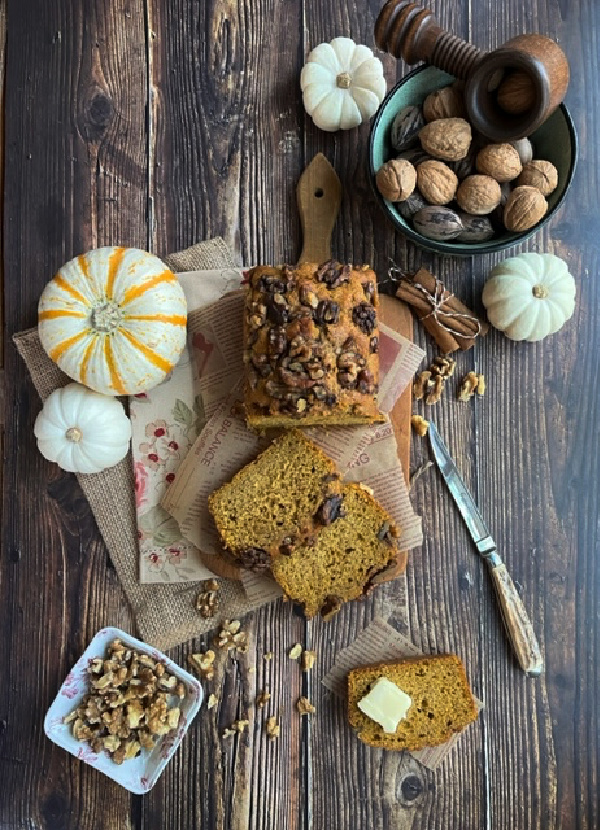 sourdough discard pumpkin bread flatlay with pumpkins bread on a wood boars, a bowl of walnuts and an antler handled knife. 