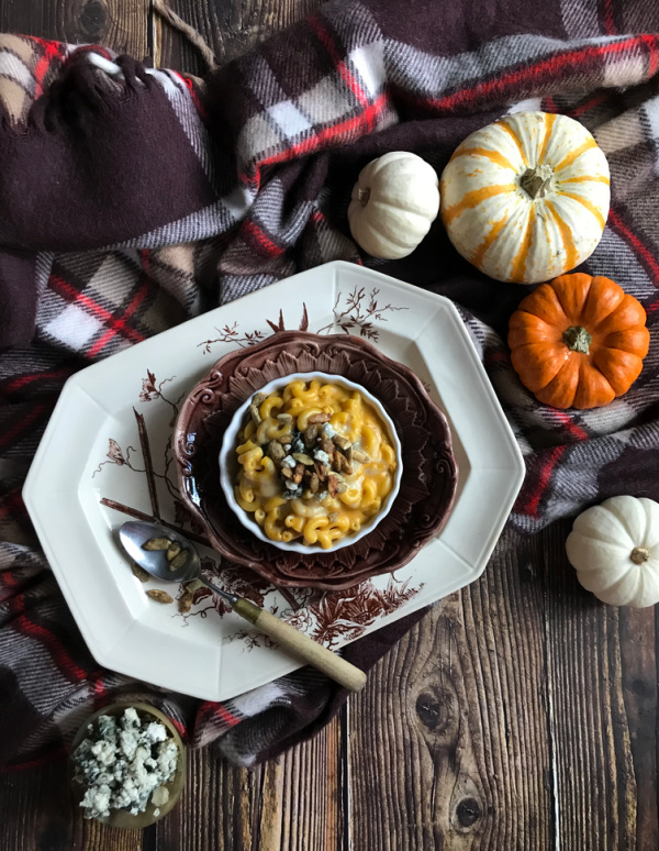 Pumpkin Mac and blue cheese in a brown ramekin, sitting on a brown and white transfer ware platter on top of a plaid blanket surrounded by orange and white pumpkins.